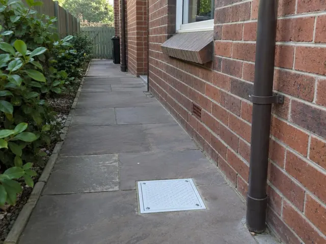 Paved side path beside a brick house with a square drain cover in place in Fairlight