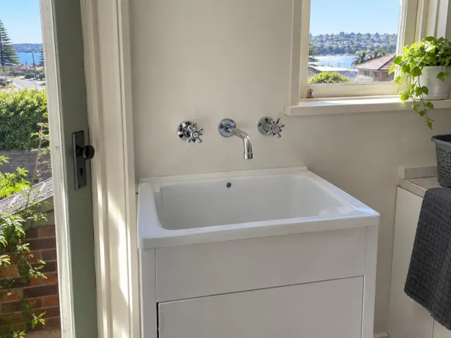 Wall-mounted tap over a white laundry sink beside a window with a water view in Manly